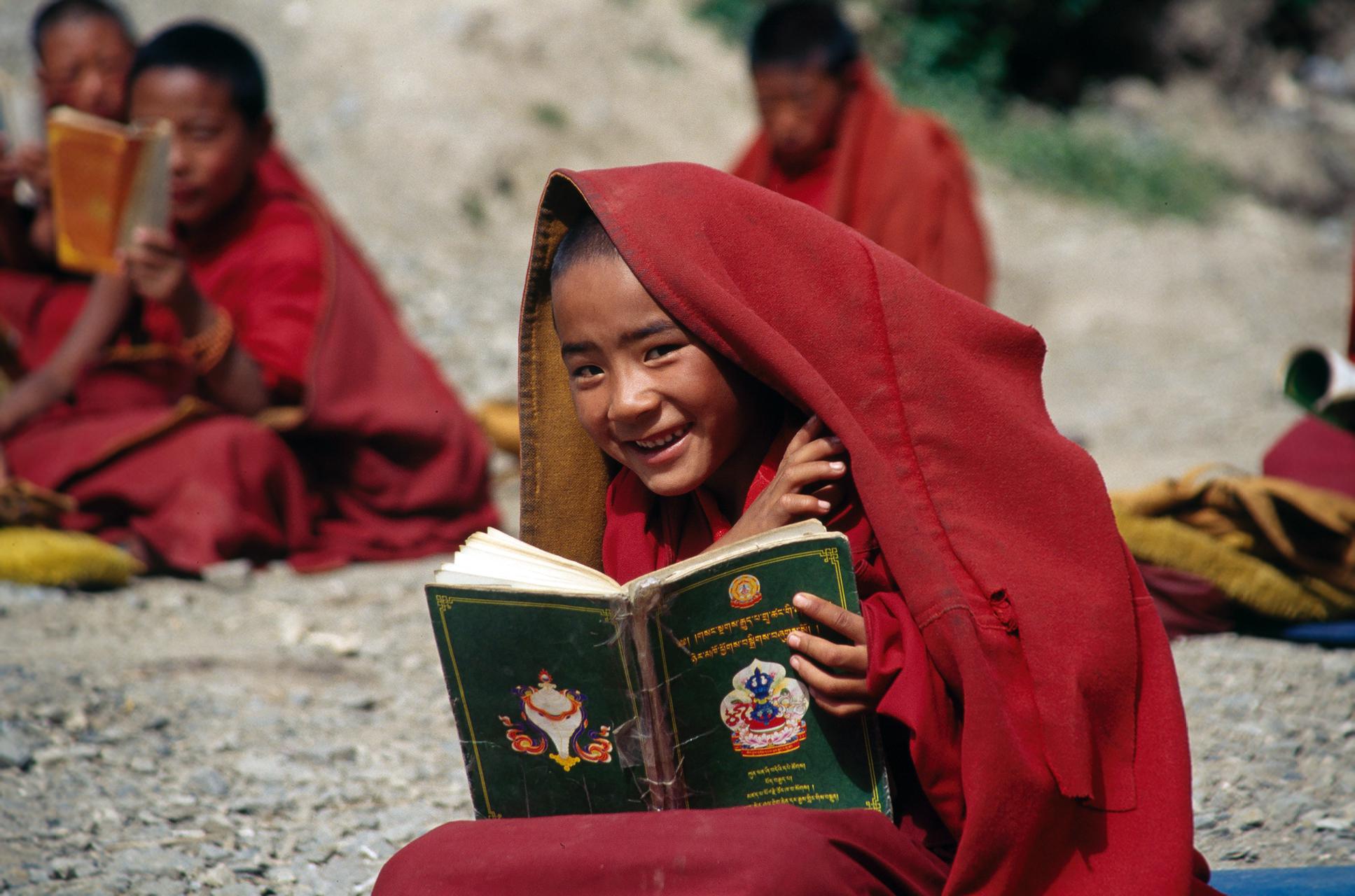 [Photo: a boy, a young Tibetan monk, reading a book with Tibetan letters]